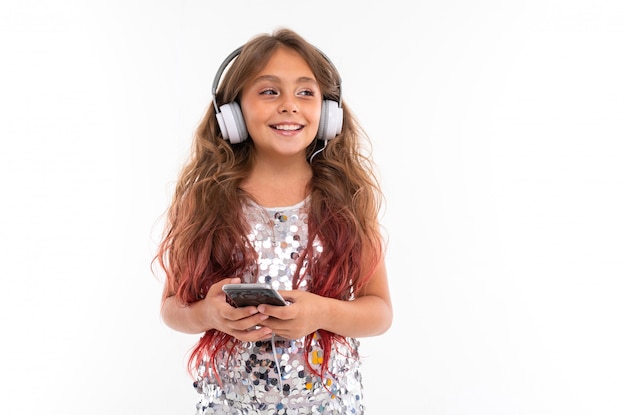 Chica en vestido brillante, con grandes auriculares blancos escuchando música y sosteniendo smartphone negro aislado