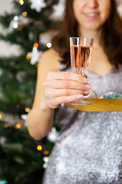 Una chica con un vestido brillante sostiene una copa de champán en el fondo de un árbol de Navidad decorado preparándose para el año nuevo y la navidad