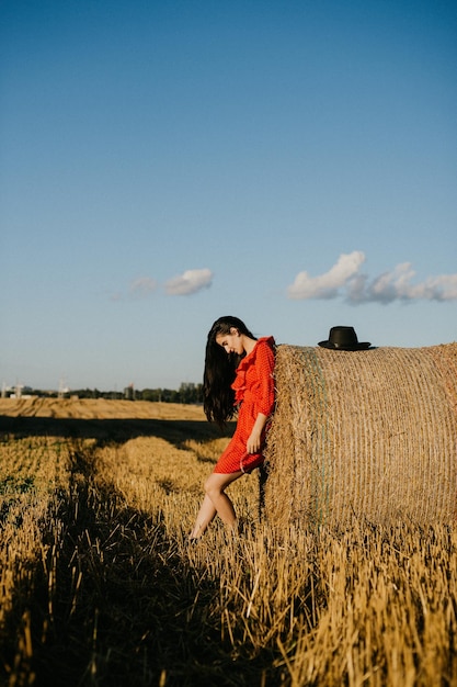 Una chica con un vestido corto rojo y zapatos negros está parada cerca de un pajar en el que hay un sombrero