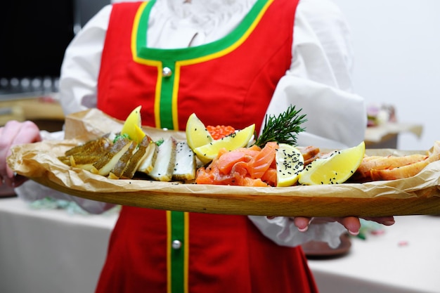 Una chica con un vestido folclórico ruso rojo sostiene una bandeja de madera con un plato de pescado en sus manos Buffet al estilo ruso