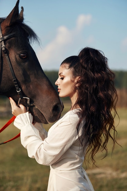 Chica en un vestido largo se encuentra cerca de un caballo, una hermosa mujer acaricia un caballo y sostiene la brida en un campo en otoño. Vida en el campo y moda, noble corcel