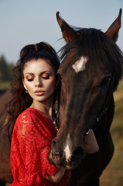 Chica con un vestido largo se encuentra cerca de un caballo, una hermosa mujer acaricia un caballo y sostiene la brida en un campo en otoño. Vida y moda en el campo, noble corcel