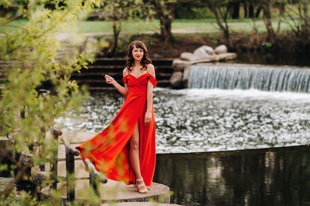 Chica con un vestido largo rojo cerca del lago en el parque al atardecer.