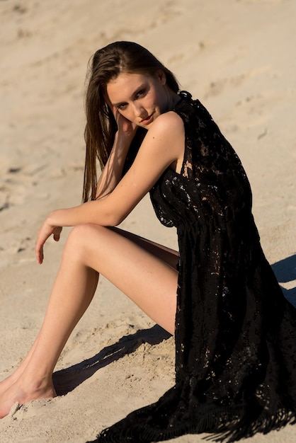 Una chica con un vestido negro se sienta en la playa en la arena.