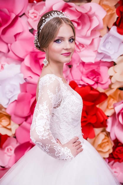 Una chica con un vestido de novia blanco con un maquillaje de espalda abierta y un peinado en forma de moño posa sobre un fondo rojo brillante de flores de papel Chica sonriendo y mirando a la cámara