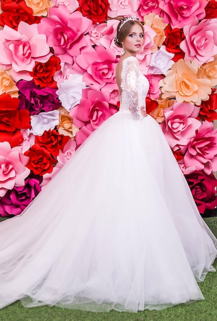 Una chica con un vestido de novia largo y blanco posa sobre un fondo rojo brillante de flores de papel