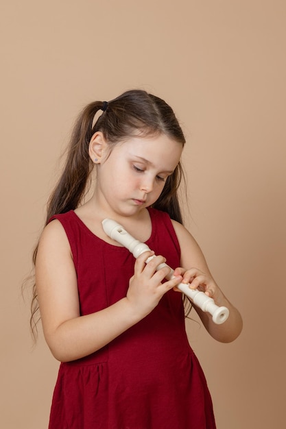 Chica en vestido rojo aprende melodía pellizcando agujeros en flauta con cabeza inclinada fondo beige Matrícula para tocar instrumentos musicales de viento de madera