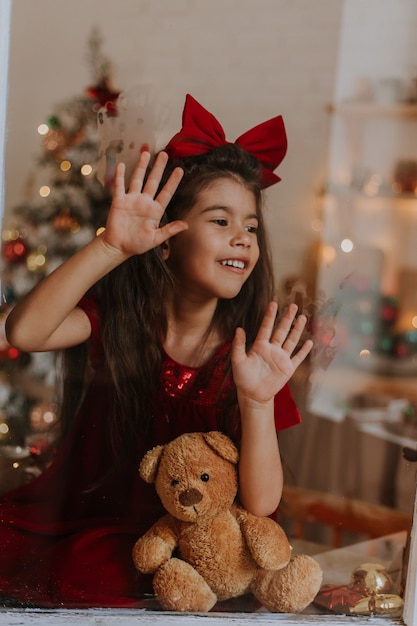 Chica con un vestido rojo con un moño en la cabeza con un oso de juguete está esperando a Santa Claus en la ventana