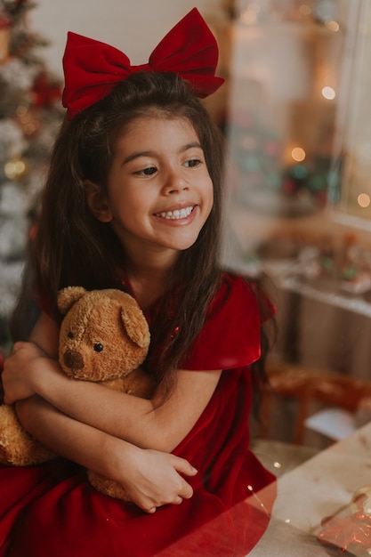 Chica con un vestido rojo con un moño en la cabeza con un oso de juguete está esperando a Santa Claus en la ventana