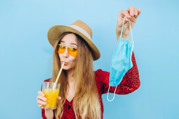 Foto chica en vestido, sombrero y gafas de sol con máscara médica en su rostro y cóctel en sus manos. vacaciones en la epidemia de coronavirus.