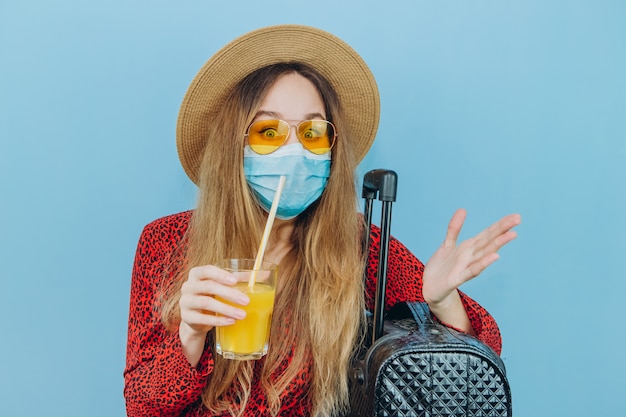 Foto chica en vestido, sombrero y gafas de sol con máscara médica en su rostro y cóctel en sus manos.