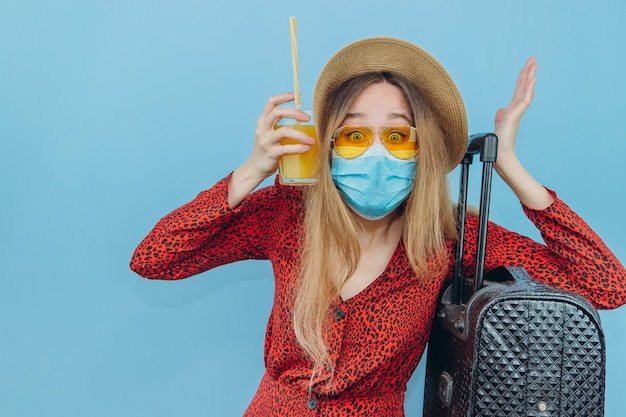 Chica en vestido, sombrero y gafas de sol con máscara médica en su rostro y cóctel en sus manos.