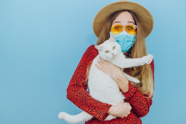 Chica en un vestido, sombrero y gafas de sol con una máscara médica en su rostro. Vacaciones en la epidemia de coronavirus. autoaislamiento con mascotas. La niña sostiene un gato blanco en sus brazos. COVID-19