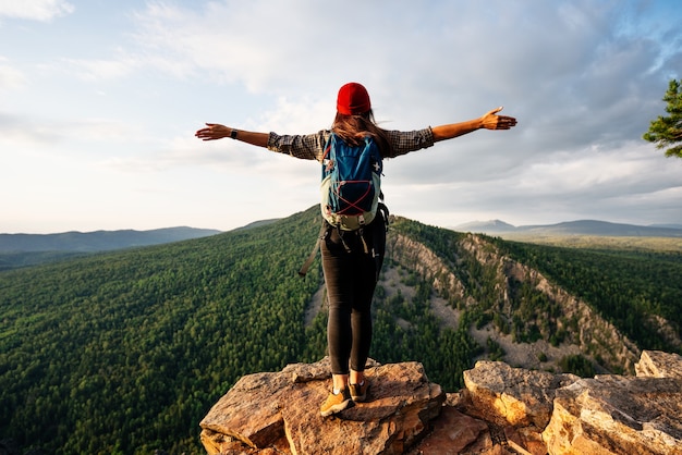 Una chica viajera con una mochila está parada en el borde de la montaña, una vista trasera. Una mujer joven con una mochila de pie al borde de un acantilado y mirando al cielo con las manos levantadas.