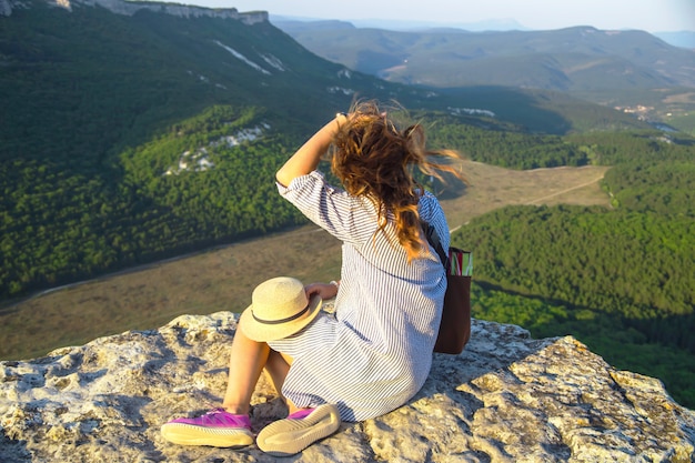 Foto una chica viajera se sienta en la cima de una montaña y sostiene su sombrero.