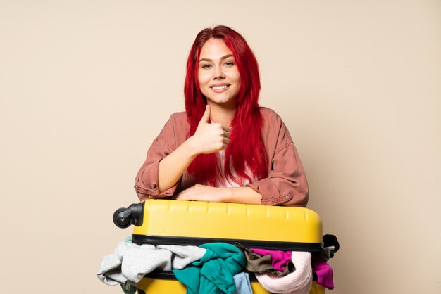 Chica de viajero con una maleta llena de ropa aislado en beige dando un gesto de pulgares arriba