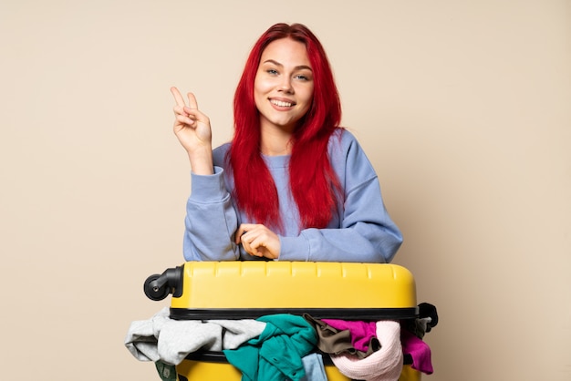 Chica de viajero con una maleta llena de ropa aislado sobre fondo beige sonriendo y mostrando el signo de la victoria