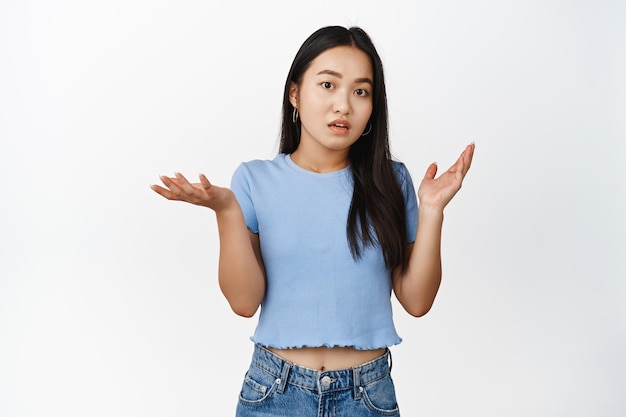 Chica vietnamita confundida encogiéndose de hombros mirando despistada no entiendo no tengo nada que decir de pie en camiseta sobre fondo blanco