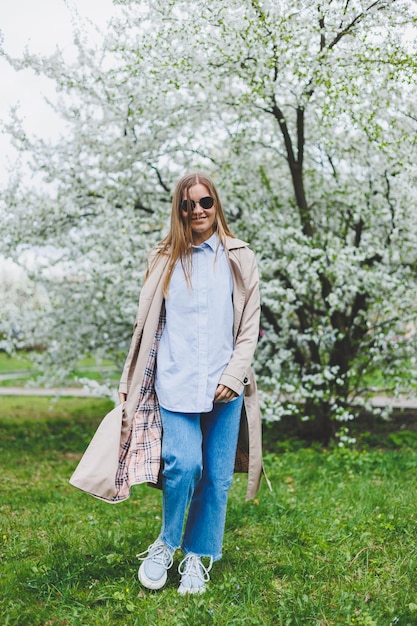 Chica de vista trasera caminando bajo un manzano floreciente en un parque natural en la ladera de una colina en Praga Elegante joven con sombrero y abrigo marrón disfrutando del jardín florido en el soleado día de primavera