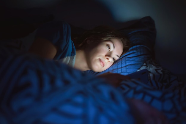 La chica yace en casa por la noche en la cama con un teléfono en la oscuridad de primer plano desenfocado