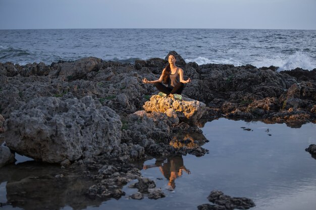 Chica yogui en postura de loto en las rocas cerca del mar por la noche