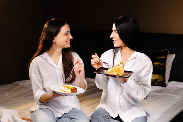 Chicas alegres comiendo pasteles sentados en el dormitorio en el apartamento