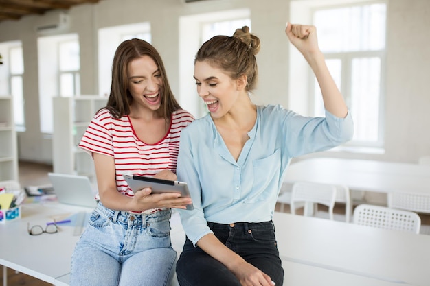 Chicas alegres sentadas en el escritorio gritando alegremente mientras usan una tableta junto con una moderna oficina vacía en el fondo