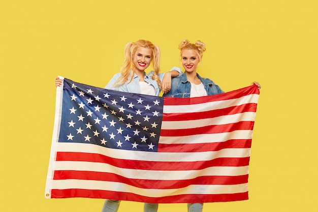 Chicas americanas. Felices mujeres jóvenes en ropa de mezclilla sosteniendo la bandera de Estados Unidos aislado sobre fondo amarillo