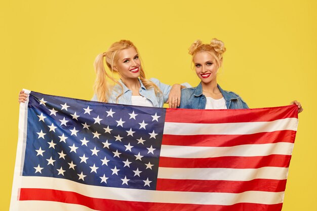 Chicas americanas. Felices mujeres jóvenes en ropa de mezclilla sosteniendo la bandera de Estados Unidos aislado sobre fondo amarillo