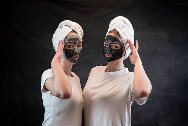 Chicas amigas en toalla blanca aplicando mascarilla facial de arcilla de barro negro aislada en negro Tratamiento antiedad cuidado de la piel