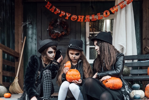 Chicas asustadizas hermanas amigas celebrando halloweenTreak or treat gameCalabazasSentado en los escalones del porcheBolsas con dulces en las manosCara aterradora maquillaje de calaveraDisfraces con estilo de brujaFiesta infantil