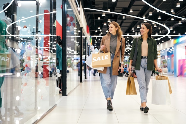 Chicas atractivas felices en trajes casuales caminando por el centro comercial y disfrutando de las compras juntos