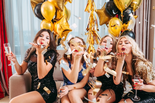 Chicas con clase divirtiéndose en la fiesta. Señoras en mini vestidos sentados en el sofá con champán, soplando confeti.