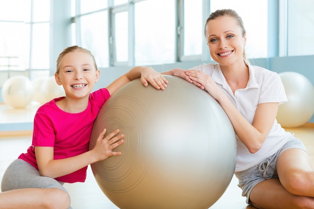 Chicas en club deportivo. Madre e hija alegres apoyándose en la pelota de fitness y sonriendo mientras están sentados en el suelo en el club deportivo