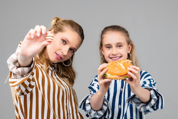 Chicas jóvenes alegres sosteniendo una hamburguesa grande en ambas manos mientras su hermana muestra una versión más pequeña