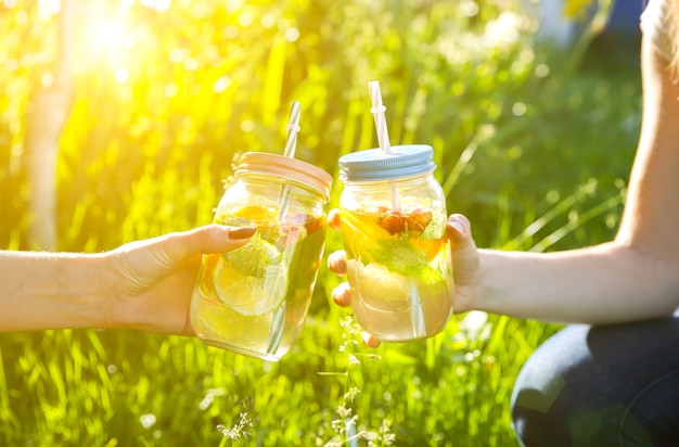 Chicas con limonada fresca en frascos con pajitas. Bebidas de verano hipster. Ecológico en la naturaleza. Limones, naranjas y bayas con menta en el vaso. Hierba verde alta al aire libre.