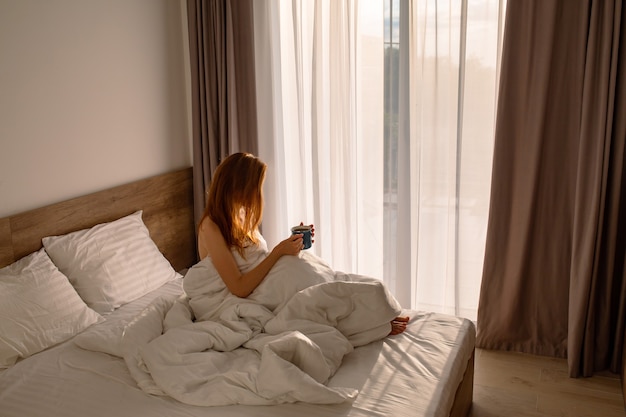 Chicas de la mañana con café al despertar a una chica en ropa de cama blanca con café