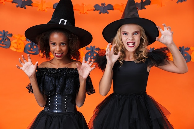 Chicas multinacionales enojadas en disfraces de halloween negros rascarse y atemorizar aislado sobre la pared de calabaza naranja