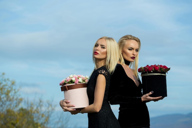 Chicas muy lindas o hermosas gemelas rubias en sexy vestido negro con flores rosas rojas y blancas en caja al aire libre sobre fondo natural con cielo azul nublado