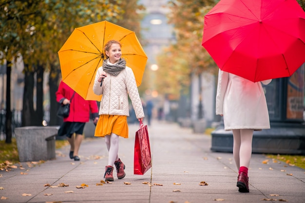 Las chicas con un paraguas rojo y amarillo se encontrarán en la calle otoñal de la ciudad. Debajo de sus pies, se cose el follaje seco caído.