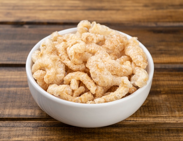 Chicharrones de cerdo frito en un recipiente blanco sobre mesa de madera.