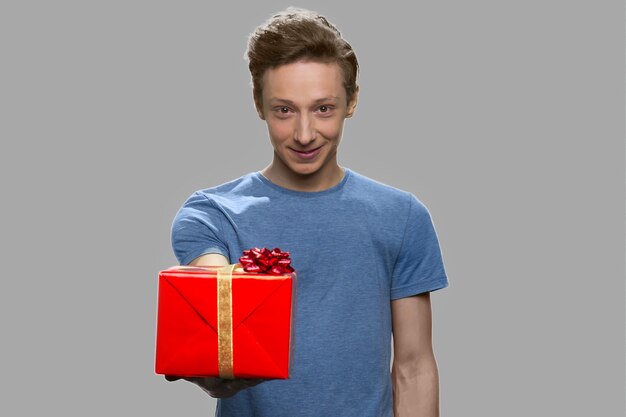 Chico adolescente dando caja de regalo a la cámara. Adolescente guapo que ofrece caja actual sobre fondo gris.