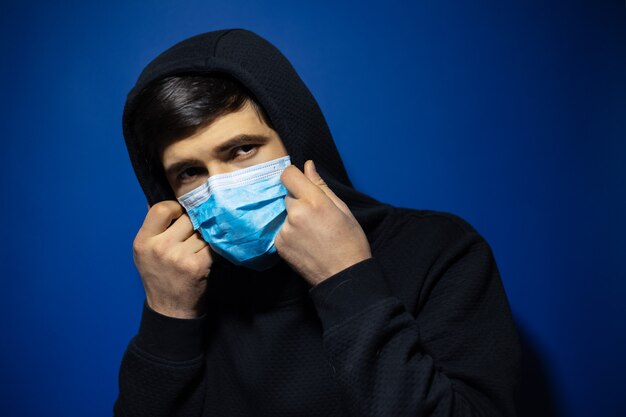 chico adolescente, con máscara médica contra la gripe, vestido con un suéter con capucha en la pared de color azul oscuro.