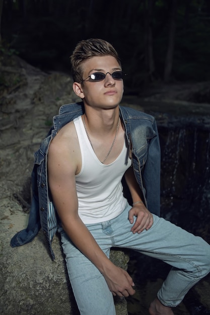 Chico adolescente de moda sexy con una camiseta blanca y gafas de sol de jeans azul, está sentado en una roca junto a una cascada en un bosque oscuro en verano