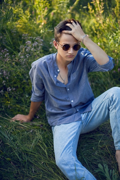 Chico adolescente sexy en gafas de sol está sentado en la hierba en la naturaleza con una camisa y pantalones vaqueros descalzo y mirando a la cámara