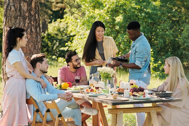 Chico africano con camisa de mezclilla y jeans blancos vertiendo vino tinto en copa de vino para una de las niñas durante la cena al aire libre junto a la mesa servida en el parque