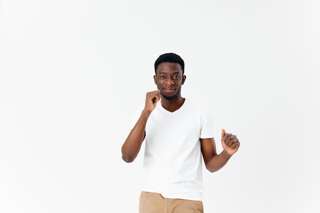 Chico africano con una camiseta blanca y pantalones beige haciendo un gesto con las manos en una vista recortada de fondo claro