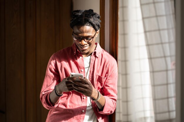 Chico africano feliz parado junto a la ventana en casa sosteniendo un teléfono celular chateando en línea usando una aplicación de citas