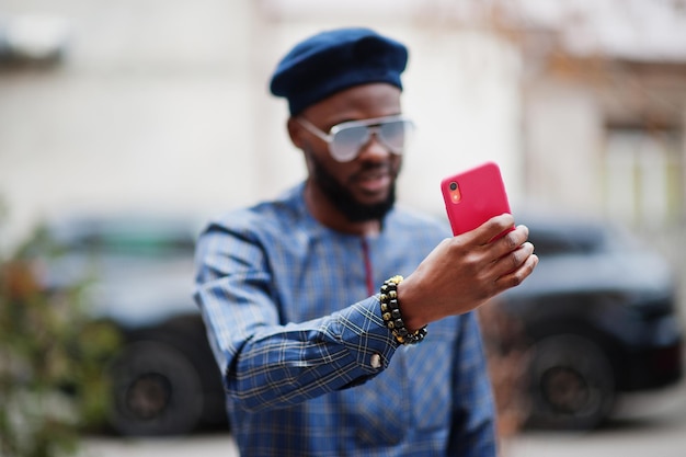 Chico africano en traje azul auténtico, anteojos y boina. Hombre nigeriano de moda cerca de coche suv negro haciendo selfie en teléfono.