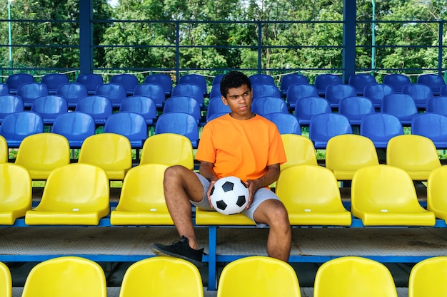 Chico afroamericano con un balón de fútbol sentado en una tribuna de fútbol vacía